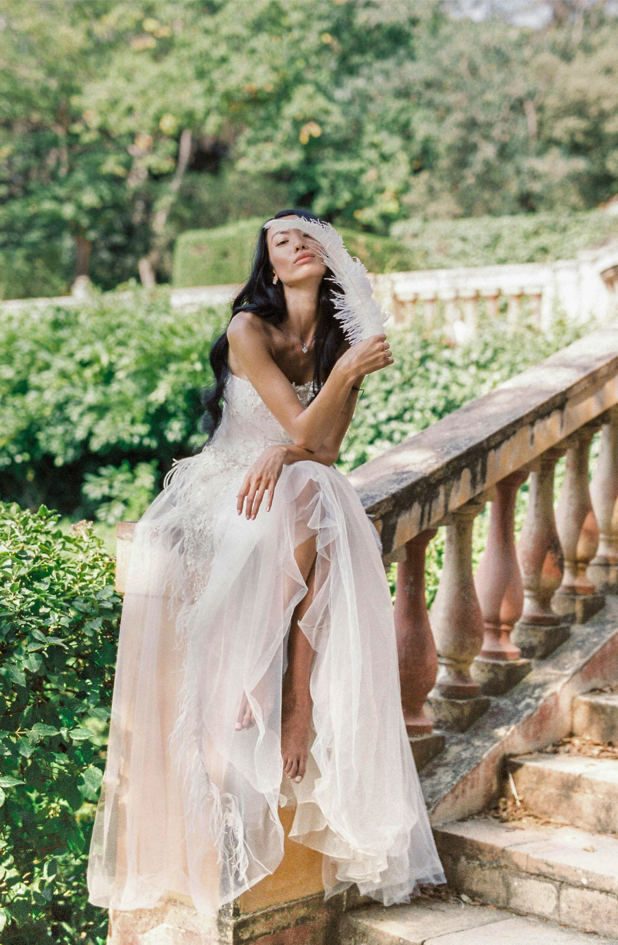 A woman in a flowing white dress sits on stone steps, holding a feathery prop, surrounded by lush greenery. The scene conveys a serene and dreamy vibe.