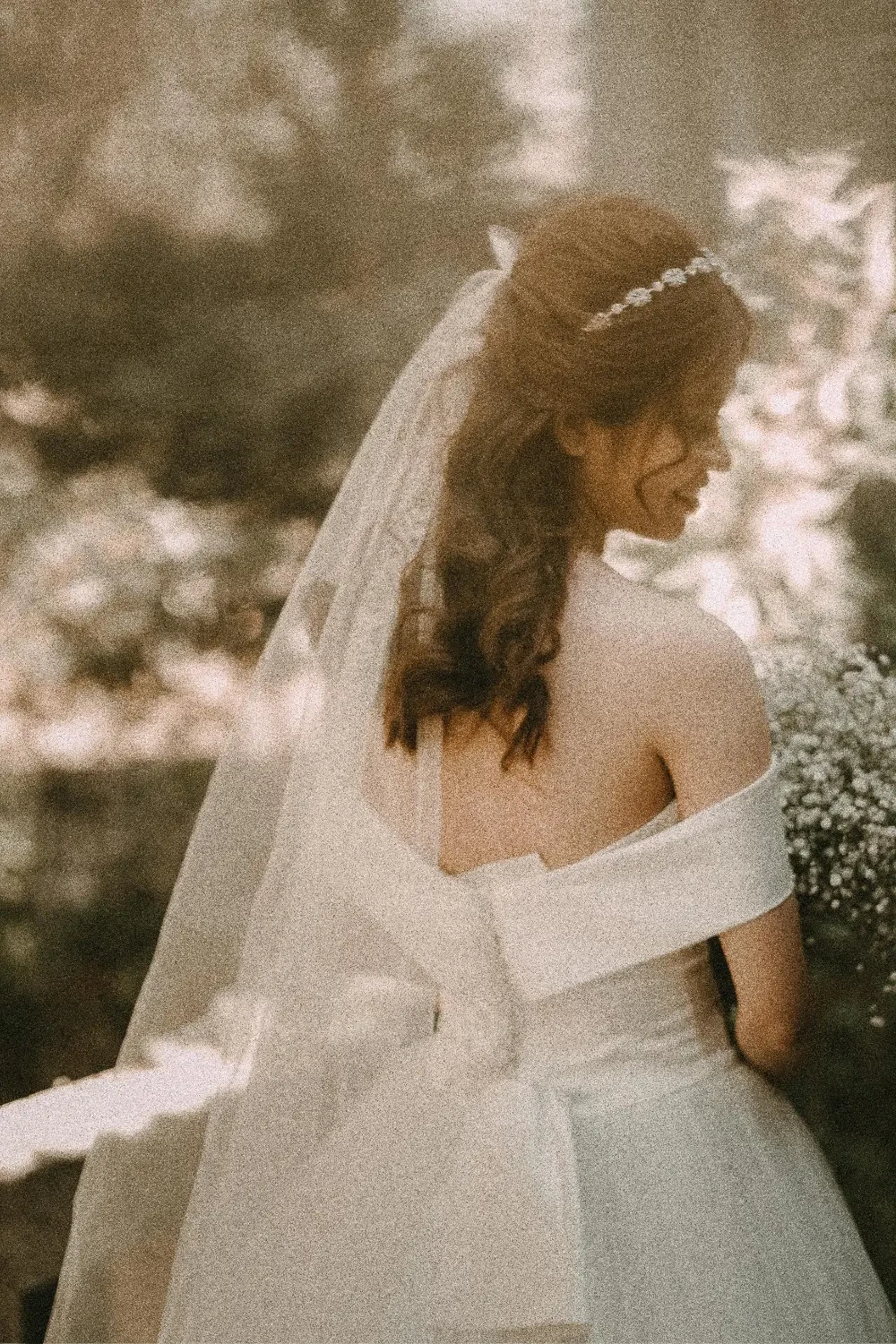 A woman in a wedding dress with a veil, seen from behind, in a soft-focus outdoor setting.