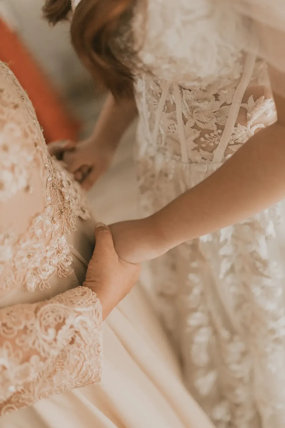 A close-up of two women holding hands, one wearing an intricately detailed wedding dress and the other in a more simple gown, with a soft background.