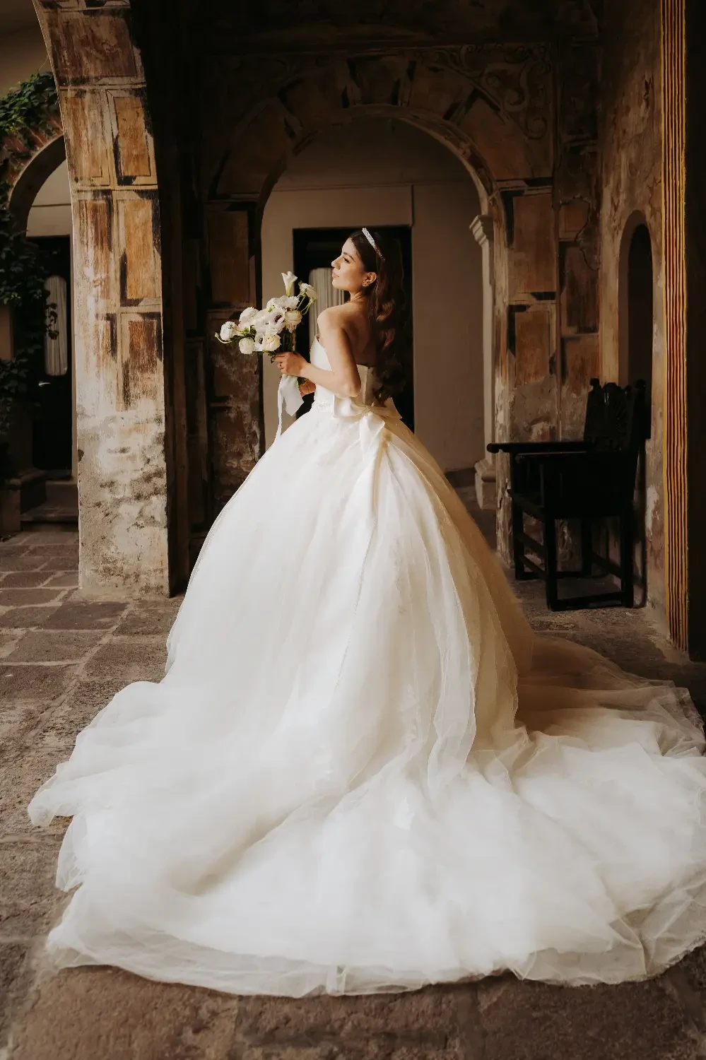 A woman in a wedding dress stands gracefully, holding a bouquet, in a picturesque indoor setting with arched architecture.