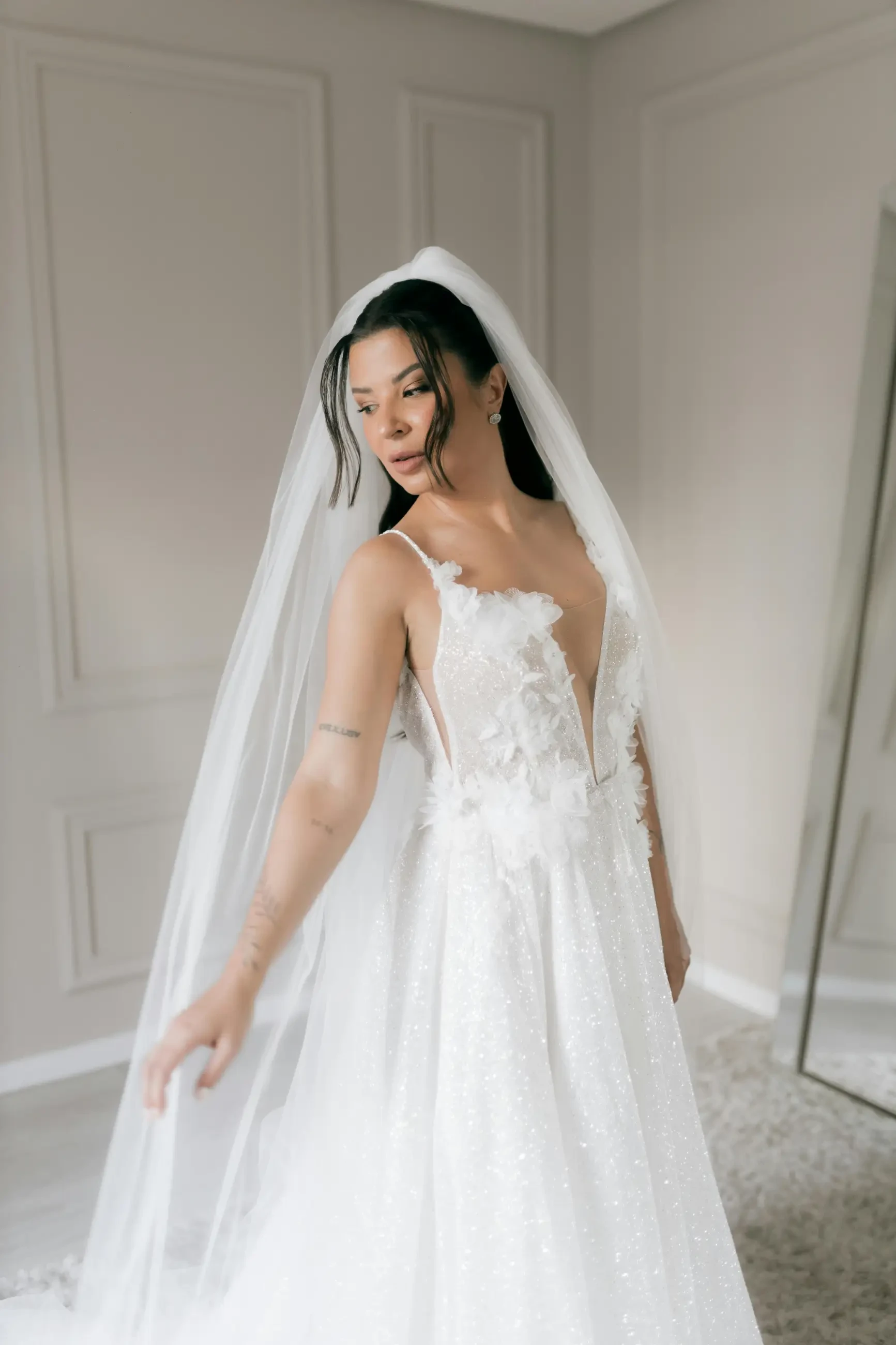 Bride in an elegant, floral lace gown with a deep V-neckline and long veil. She poses gracefully in a serene, softly lit room, exuding elegance.