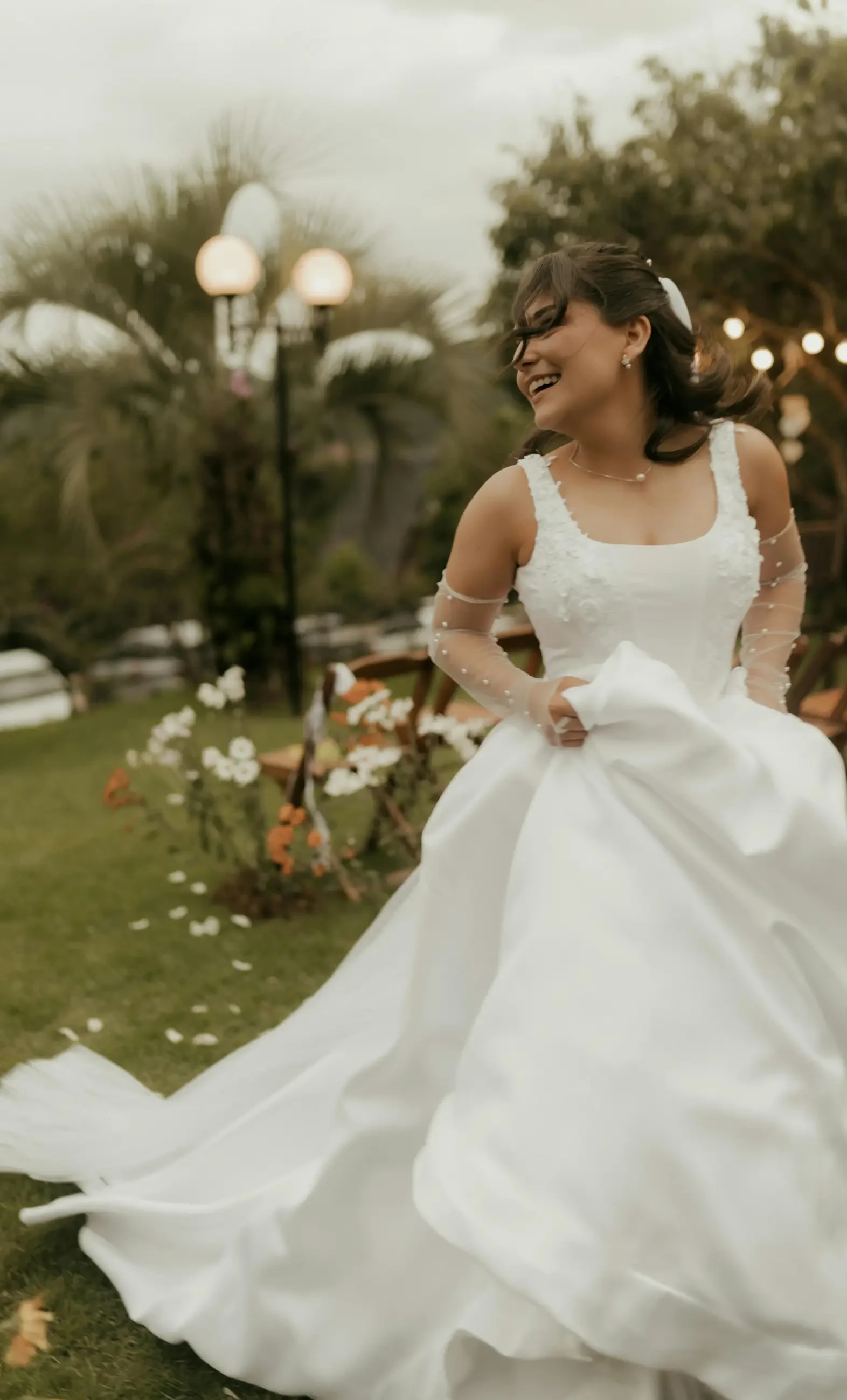 A smiling woman wearing a white wedding dress, twirling in a garden filled with flowers and greenery. Soft lighting creates a warm atmosphere.
