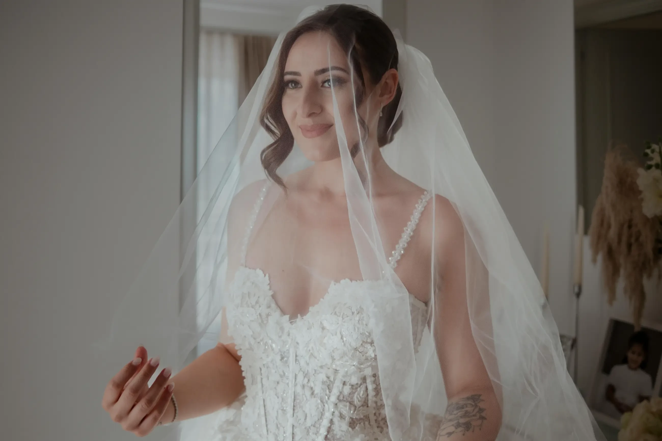 A bride in a lace wedding dress and veil smiles softly, her fingers gently holding the veil. The room is softly lit, conveying a warm, joyful ambiance.