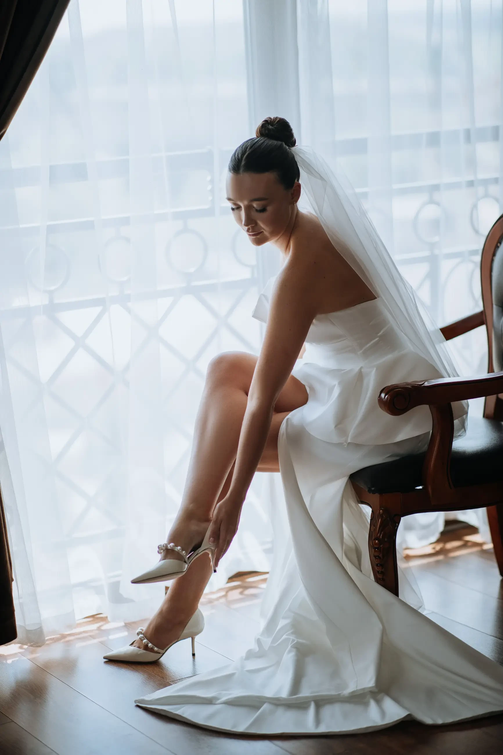 A bride sitting on a chair while putting on her high-heeled shoes, with a sheer curtain in the background.