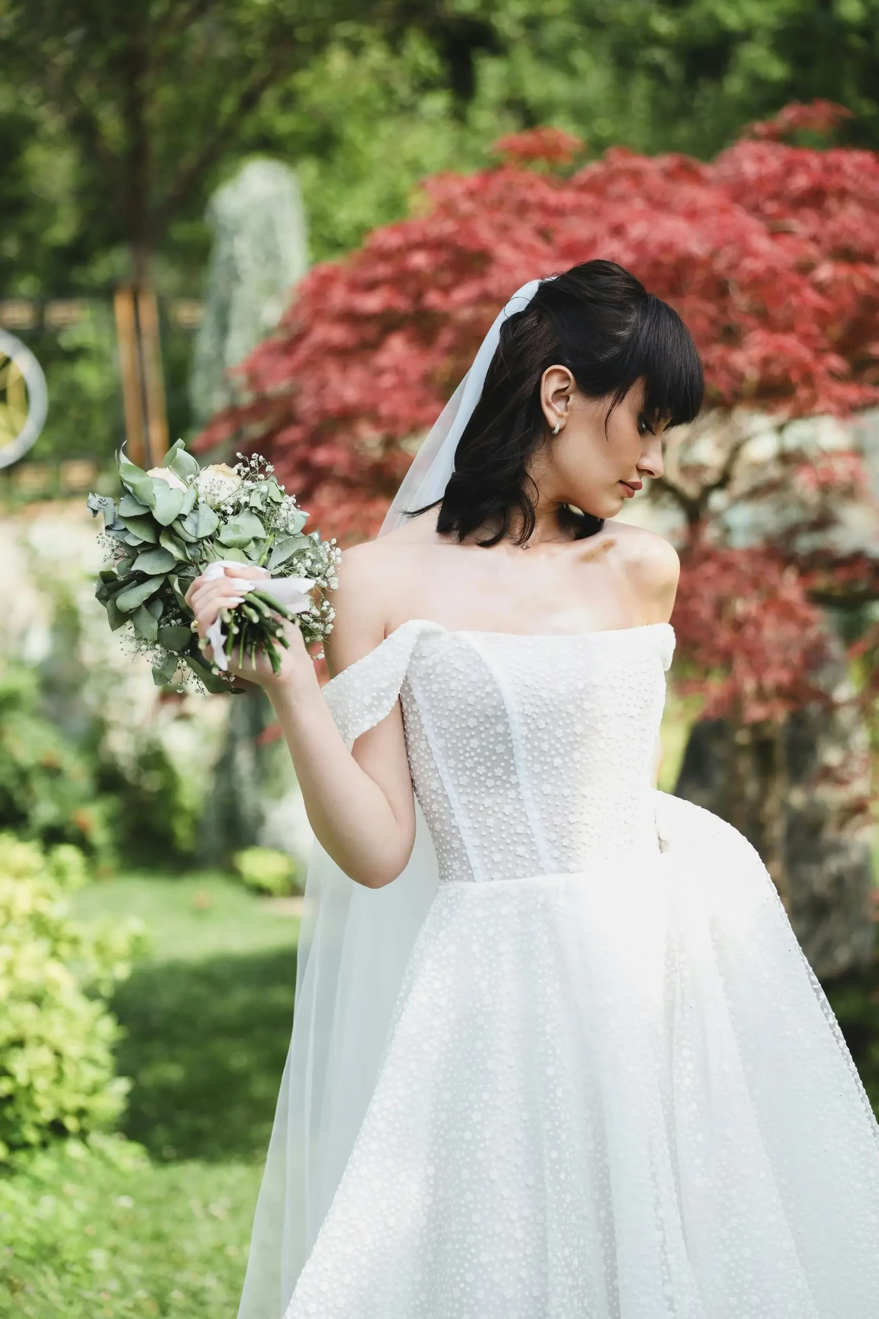 A bride in an elegant off-shoulder wedding dress, holding a bouquet of greenery, standing in a lush garden with trees and plants around her.