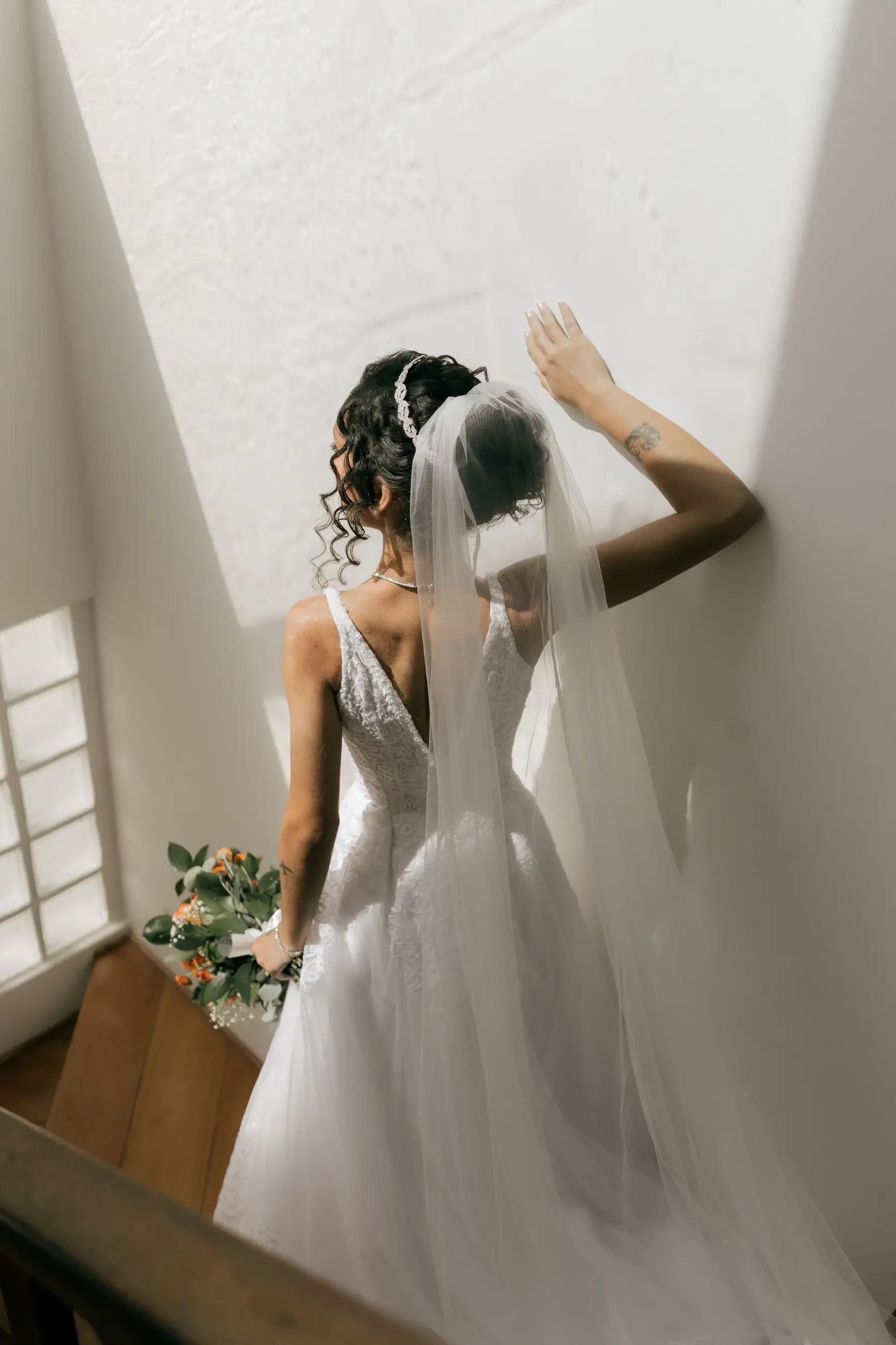 A bride holding a bouquet, wearing a white wedding dress and veil, standing against a wall with sunlight streaming in.