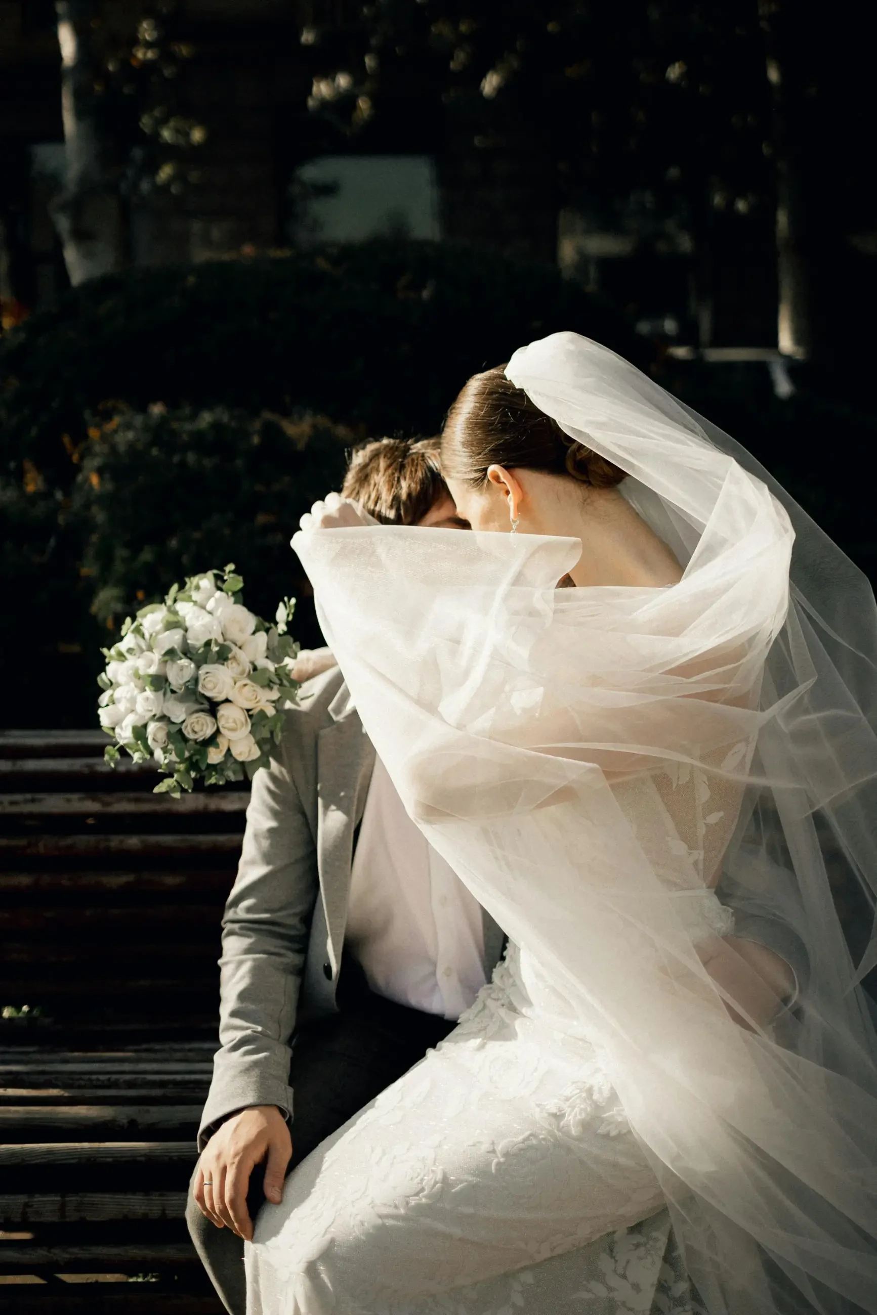 Bride and groom sitting on a bench, with her veil elegantly flowing in the breeze. The bride holds a bouquet of white roses, creating a romantic scene.