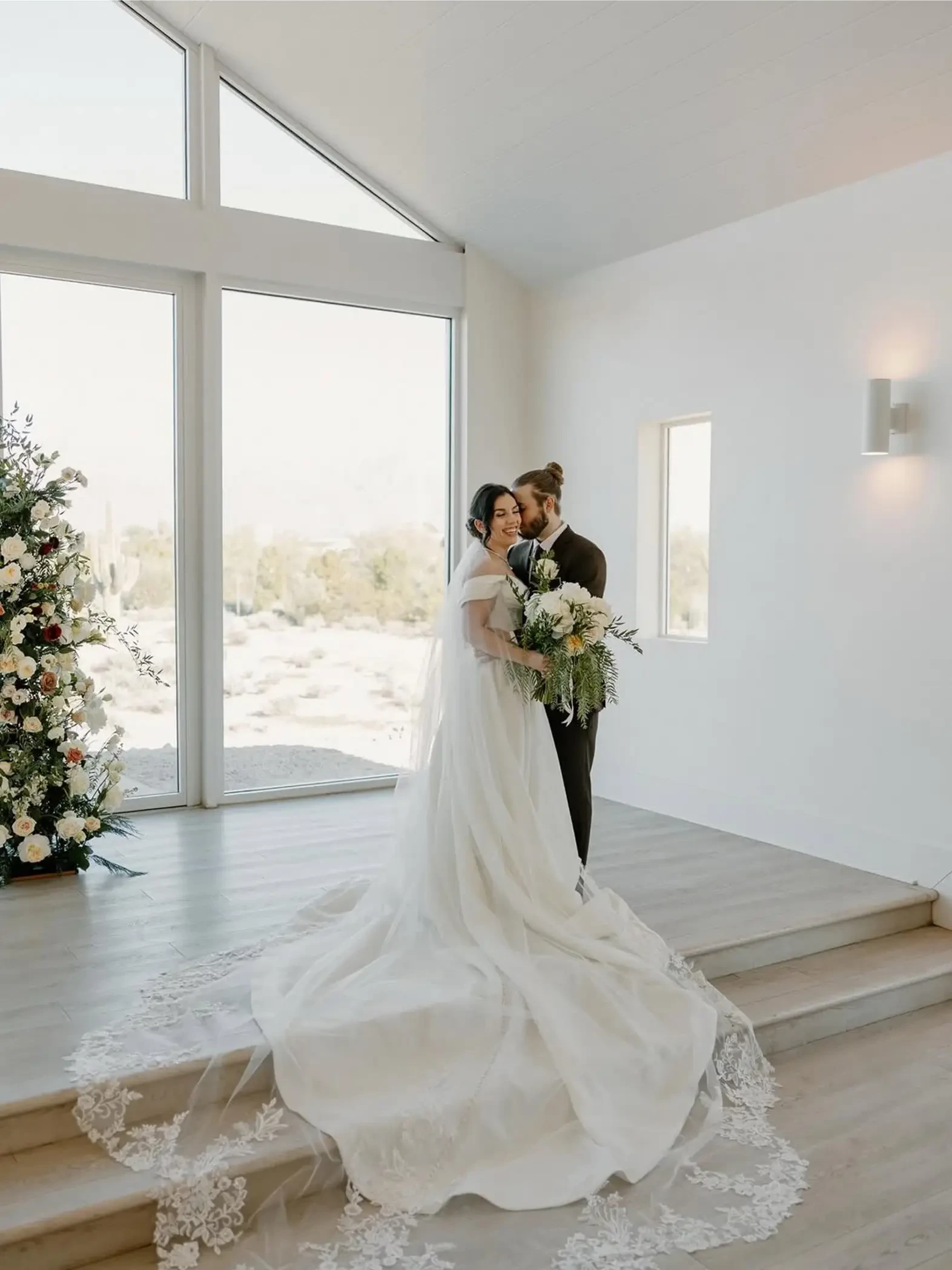 A bride and groom embrace in a bright, modern venue decorated with floral arrangements. The bride is wearing a white gown with intricate lace details, while the groom is dressed in a dark suit. Natural light streams through large windows, creating an elegant atmosphere.